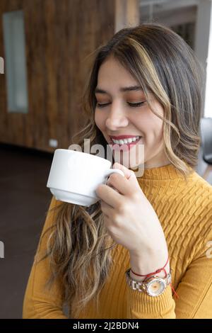 femme latine avec de longs cheveux blonds souriant, tenant une tasse avec boisson chaude, style de vie, maquillage et café petit déjeuner Banque D'Images