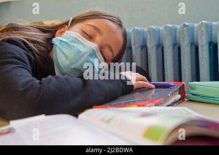 École primaire pendant l'épidémie de coronavirus Covid-19 : écolière avec masque facial endormi dans une salle de classe Banque D'Images