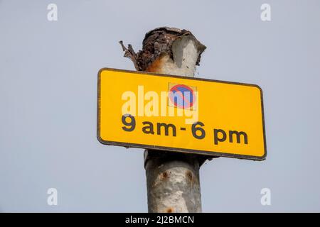 Petit panneau rectangulaire de restriction de stationnement noir sur jaune avec symbole rouge et bleu pour les heures de 9am à 6pm monté sur un poteau métallique très corrodé Banque D'Images