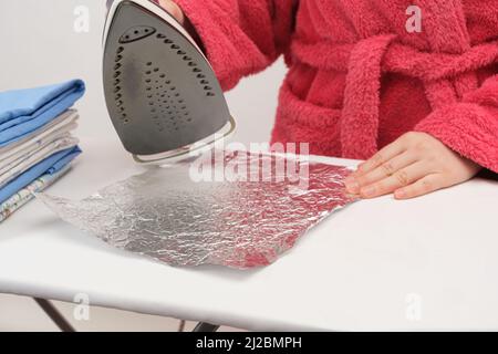 La femme au foyer nettoie la semelle du fer à repasser du dépôt avec du papier d'aluminium, repassant le papier d'aluminium sur la planche à repasser en gros plan. Banque D'Images