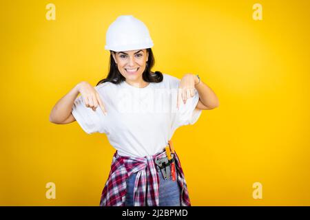 Jeune femme caucasienne portant un casque et des vêtements de construction sur fond jaune isolé surpris, regardant vers le bas et pointant vers le bas avec les doigts et r Banque D'Images
