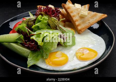 Petit déjeuner. Œufs frits avec pain grillé, beurre et légumes, concombres et tomates. Sur une plaque sombre, sur un fond sombre Banque D'Images