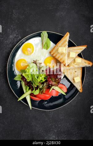 Petit déjeuner. Œufs frits avec pain grillé, beurre et légumes, concombres et tomates. Sur une plaque sombre, sur un fond sombre Banque D'Images