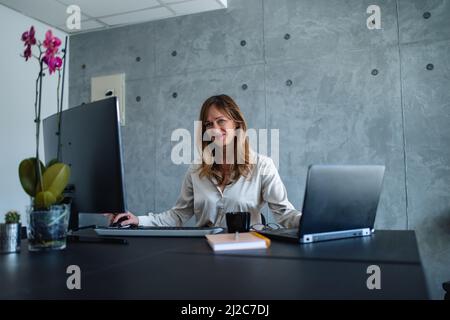 Femmes âgées de race blanche sur ordinateur portable et de bureau en même temps. Elle regarde la caméra. Banque D'Images