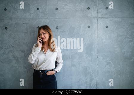 Une femme de race blanche pose pour la caméra devant le mur gris et a un appel téléphonique. Banque D'Images