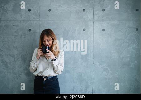 Une jolie femme est debout devant le fond gris alors qu'elle tient une tasse de boisson avec ses deux mains. Elle regarde vers le bas. Banque D'Images