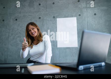 La femme souriante montre le pouce levé sur la main droite dans le bureau et la présentation correcte. Un livre blanc est suspendu sur le mur gris derrière elle. Banque D'Images