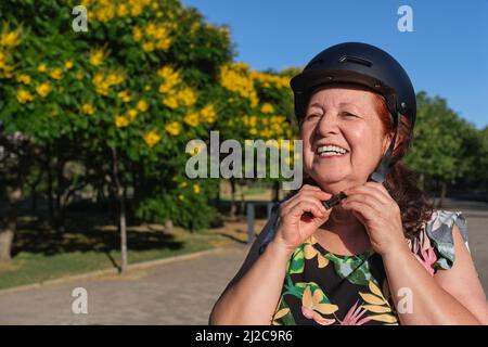 Heureuse femme hispanique mûre en train de rire en mettant un casque de sport dans un parc de la ville. Concepts de vie urbaine active, de bien-être et de sécurité routière. Banque D'Images