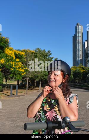 Femme hispanique mûre souriante mettant un casque pour monter à bord de son scooter électrique dans un parc de la ville. Concepts de mobilité électrique verte, urba actif Banque D'Images
