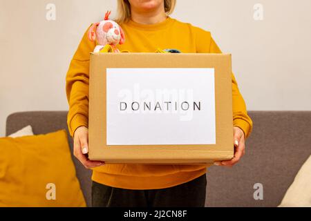 Jeune femme assise sur un canapé préparant le colis pour l'envoi à l'homme dans le besoin. Fille avec grand coeur de la sorte met les vêtements utilisés nouveau port et l'ours doux jouet dedans Banque D'Images