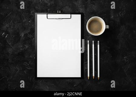 Photo de presse-papiers avec papier vierge, tasse à café et crayons sur fond de plâtre noir. Concept de bureau. Banque D'Images