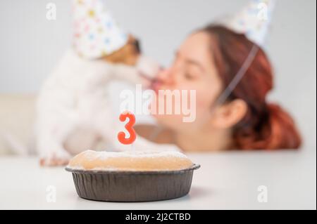 Femme caucasienne et Jack russell terrier en calottes de vacances regarder le gâteau avec une bougie. Le chien et le propriétaire célèbrent le troisième anniversaire. Banque D'Images