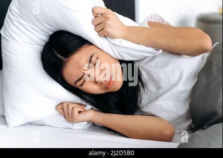 La jeune femme asiatique, couchée au lit à la maison, couvre ses oreilles avec un oreiller, tente de dormir, se ferme du bruit, a un mauvais rêve, rêve agité. Concept sommeil et repos Banque D'Images