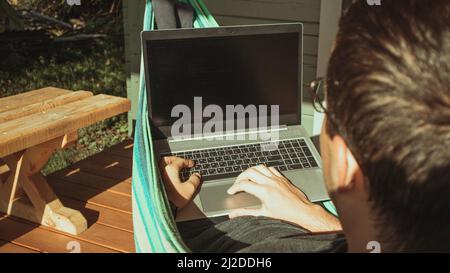 Un endroit confortable pour travailler à la maison. Un homme est assis et travaille dans un hamac près d'une maison de campagne par une journée ensoleillée Banque D'Images