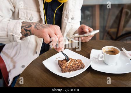 Gros plan d'un homme méconnaissable mangeant un gâteau et buvant un café tout en vérifiant son téléphone cellulaire Banque D'Images