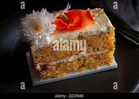 Un morceau de gâteau de carotte avec une fleur sur une assiette noire en lumière basse et avec une fourchette sur le côté. Arrière-plan noir Banque D'Images