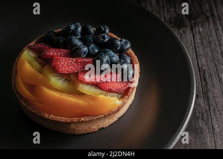 tarte fine à la pâtisserie française avec mûres de fraises, kiwis et pêches sur le dessus et sur une assiette sombre sur une table en bois rustique, photographie de nourriture sombre Banque D'Images