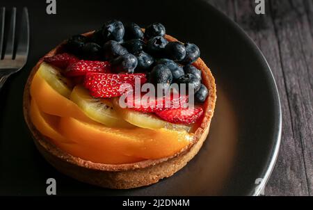 tarte fine à la pâtisserie française avec mûres de fraises, kiwis et pêches sur le dessus et sur une assiette sombre sur une table en bois rustique, photographie de nourriture sombre Banque D'Images