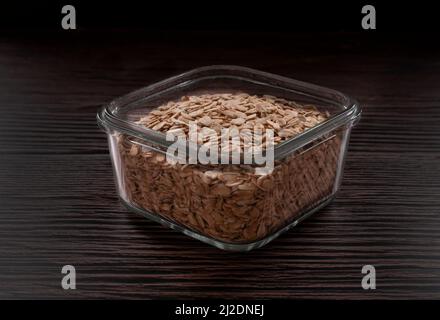 Flocons d'avoine crus dans un récipient en verre carré sur une table en bois sombre, photographie de nourriture sombre avec fond noir horizontal. Banque D'Images