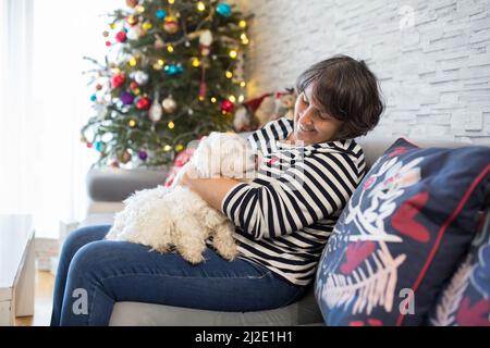 Femme d'âge moyen, tenant le chien maltais à la maison devant l'arbre de Noël, en hiver Banque D'Images