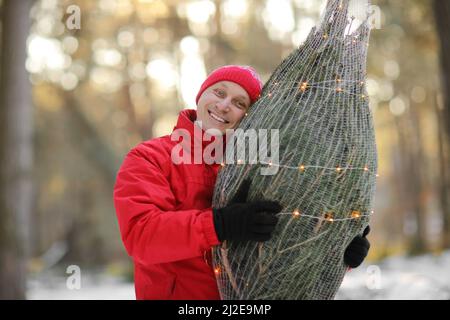 homme souriant portant un arbre de noël fraîchement coupé dans la forêt. Le jeune bûcheron porte un sapin sur son épaule dans les bois. Comportement irresponsable t Banque D'Images