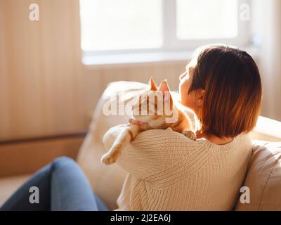 Une jeune femme asiatique porte un chandail chaud reposant avec un chat tabby sur un canapé à la maison un jour d'automne. Photo intérieure d'une femme incroyable tenant un animal de compagnie au gingembre. Heure de sommeil matinale à la maison. Mise au point douce. Banque D'Images