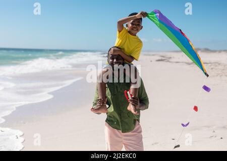 Portrait de l'homme afro-américain portant son fils avec cerf-volant sur les épaules à la plage pendant la journée ensoleillée Banque D'Images