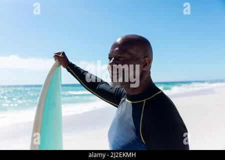 Homme senior chauve d'amérique africaine souriant tenant une planche de surf, regardant la plage par beau temps Banque D'Images