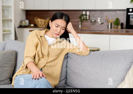 La jeune femme asiatique malheureuse assise sur un canapé, couverte de front avec la main et la frustration de sentir, la femme orientale fatiguée et déprimée seule à la maison, souffrant de maux de tête, se sent mal Banque D'Images