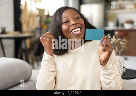 Bonne jeune femme souriant avec plaisir, assis au canapé avec une carte de crédit et regardant l'appareil photo. Bonne fille biraciale montrant carte de crédit en plastique. Concept d'achat en ligne Banque D'Images