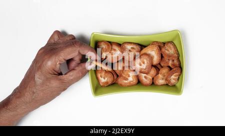 Biscuit en forme de cœur dans un bol carré vert, avec une main ramassant le biscuit, isolé sur un fond blanc Banque D'Images