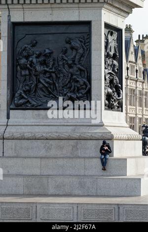 Un homme seul assis sur le socle de Nelsons Column à l'aide d'un téléphone portable Trafalgar Square, centre de Londres, Angleterre, Royaume-Uni Banque D'Images