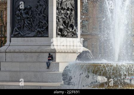 Un homme seul assis sur le socle de Nelsons Column à l'aide d'un téléphone portable Trafalgar Square, centre de Londres, Angleterre, Royaume-Uni Banque D'Images