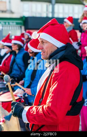 Vue rapprochée sur le côté d'un homme d'âge moyen vêtu d'une veste rouge et d'un chapeau de Père Noël, jouant le tambour dans un groupe de percussions à l'extérieur par temps froid. Banque D'Images