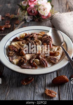 Bol de petit déjeuner avec noix de pécan grillées, dattes, bananes, pommes et cannelle sur fond de table en bois. Image verticale avec espace de copie Banque D'Images