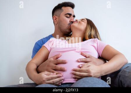 couple hétérosexuel caucasien enceinte dans le lit embrassant et embrassant le ventre, l'homme embrasse la femme enceinte sur la joue assis sur le lit avec le mur blanc dedans Banque D'Images