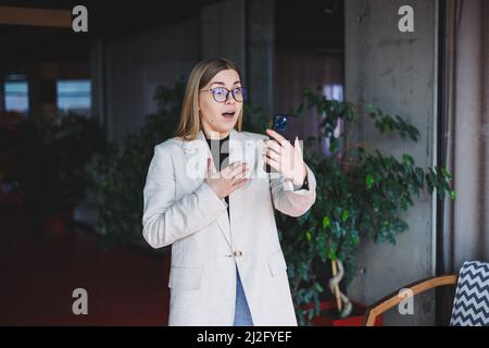 Une belle jeune femme dans une veste beige et des lunettes a lu les nouvelles sur un téléphone mobile et a été choquée par ce qu'elle a lu. Banque D'Images