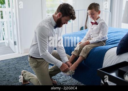 Il obtient son style de papa. Coupe courte d'un jeune beau père aidant son adorable fils à mettre ses chaussures à la maison. Banque D'Images