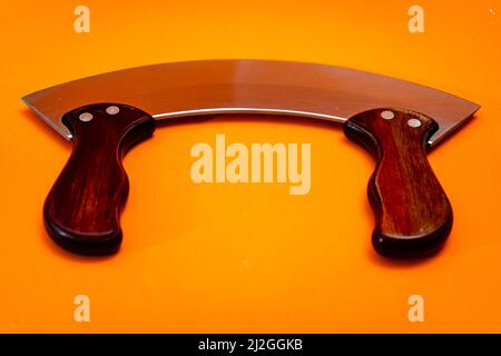 Couteau Mezzaluna avec lame en acier inoxydable et poignées en bois isolées sur fond orange. Couper les ustensiles de cuisine Banque D'Images