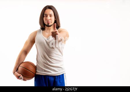 Sportif musclé joueur de basket-ball caucasien montrant les pouces jusqu'à la caméra tout en tenant le ballon de basket-ball dans son bras. Studio moyen tourné sur fond blanc. Photo de haute qualité Banque D'Images