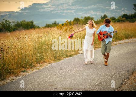 Un jeune marié joue de la guitare pour sa mariée pendant qu'ils marchent sur la route un beau jour dans la nature. Relation, lune de miel, nature Banque D'Images