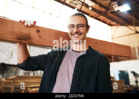La menuiserie a toujours été ma passion. Portrait d'un beau jeune charpentier portant une planche de bois sur ses épaules à l'intérieur d'un atelier. Banque D'Images