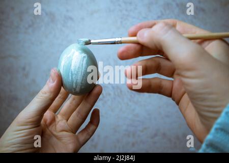 Main tenant un pinceau et peignant un oeuf. Un processus de peinture des oeufs de Pâques avec de la peinture gris-bleu. Pinceau dans la main de femme gros plan. Tradition et Banque D'Images