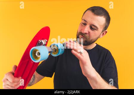 Photo studio moyen d'un homme européen d'âge moyen tenant son mini-skateboard rouge et touchant l'une de ses roues sur fond jaune. Photo de haute qualité Banque D'Images