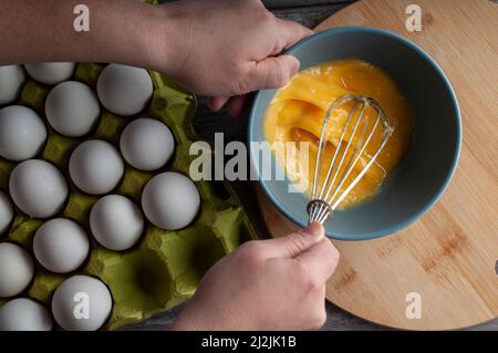 œufs brouillés crus dans un bol bleu sur une planche à découper ronde en bois et les mains mélangeant les œufs, Banque D'Images