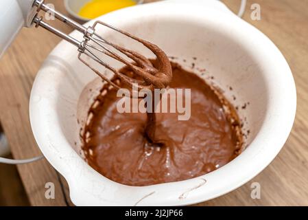 Batteur à main avec un fouet surélevé au-dessus du bol, avec de la pâte crue, la pâte est dans un état liquide épais brun. Banque D'Images