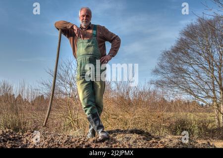 Un fermier dans des salopettes vertes et des bottes en caoutchouc est debout sur un tas de dung et s'est penché contre sa fourche de dung. Banque D'Images