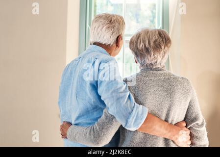 Souvenirs derrière eux, l'avenir devant eux. Vue arrière d'un couple senior affectueux qui regarde par une fenêtre ensemble à la maison. Banque D'Images