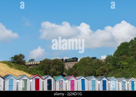 Maisons de plage colorées. Rangée de cabanes de plage multicolores avec train à vapeur d'époque sur viaduc de pierre contre ciel bleu. Banque D'Images