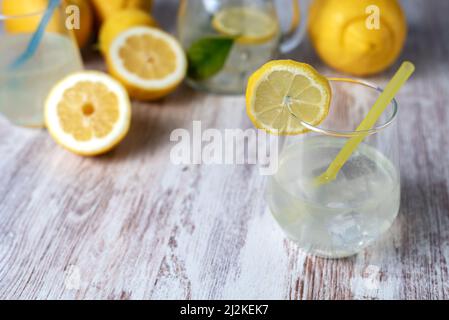 Verre avec limonade et glace, avec une tranche de citron sur son bord, en arrière-plan un pichet et quelques citrons hors foyer. Banque D'Images